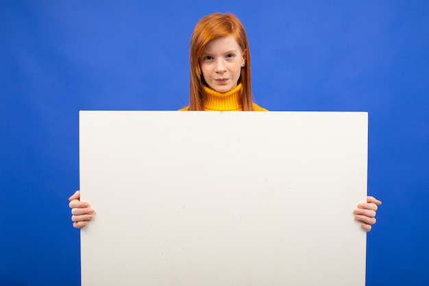 Charmante adolescente rousse européenne tenant une affiche en papier vierge pour la publicité sur bleu