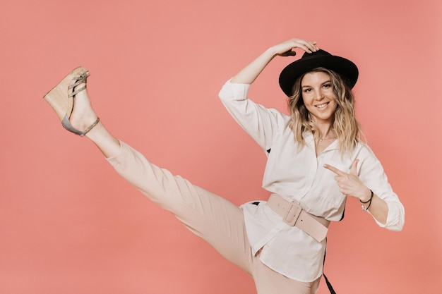 Charmante blonde souriante dans un chapeau avec un chemisier blanc à bords et un pantalon rose clair faisant un coup de pied avec elle