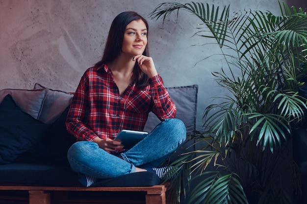 Charmante brune dans une chemise en flanelle et un jean assis sur un canapé