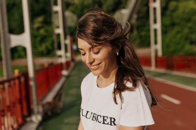 Charmante dame adorable aux cheveux longs portant un tshirt blanc souriant avec les yeux fermés au soleil le matin