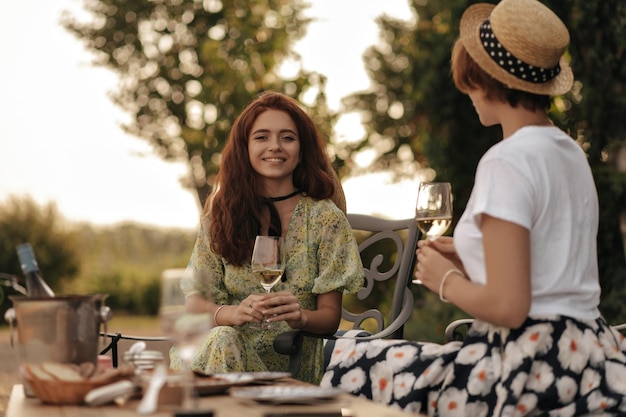 Charmante dame aux cheveux roux en robe d'été élégante souriant tenant un verre de vin et assis sur une chaise avec un ami moderne en plein air Les filles ont pique-nique