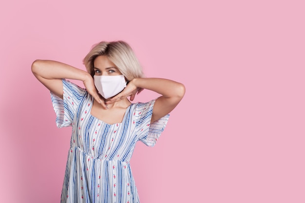 Charmante dame blonde avec un masque médical portant une robe pose sur un mur de studio rose avec espace libre