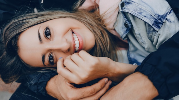 Charmante dame de race blanche avec un sourire joyeux à pleines dents est allongée sur son petit ami embrassé par lui