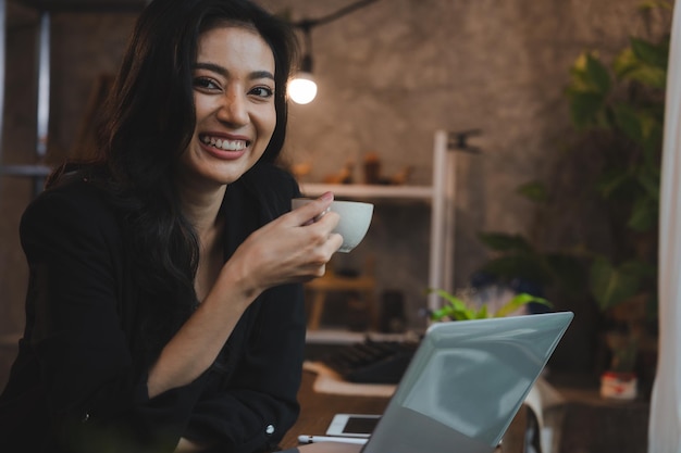 Charmante femme d'affaires asiatique intelligente entrepreneur utilise un ordinateur portable pour travailler entreprise de formation séminaire de développement de carrière s'asseoir à table tenir une tasse à emporter boisson chaude dans un loft bureau ou un café mode de vie de travail indépendant