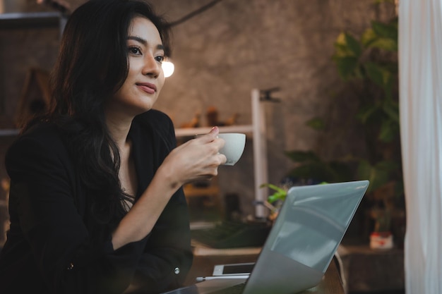 Charmante femme d'affaires asiatique intelligente entrepreneur utilise un ordinateur portable pour travailler entreprise de formation séminaire de développement de carrière s'asseoir à table tenir une tasse à emporter boisson chaude dans un loft bureau ou un café mode de vie de travail indépendant