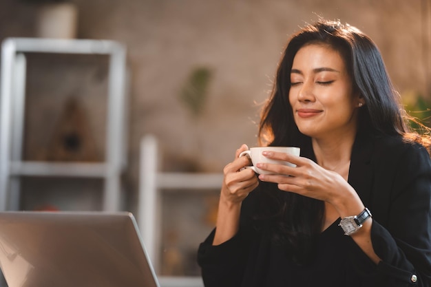 Charmante femme d'affaires asiatique intelligente entrepreneur utilise un ordinateur portable pour travailler entreprise de formation séminaire de développement de carrière s'asseoir à table tenir une tasse à emporter boisson chaude dans un loft bureau ou un café mode de vie de travail indépendant