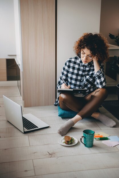 Photo charmante femme d'affaires caucasienne écrire quelque chose dans le livre alors qu'il était assis sur le sol avec un ordinateur portable et une tasse de thé