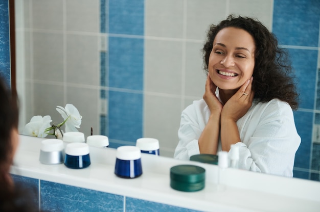 Charmante femme afro-américaine d'âge moyen sourit en regardant son reflet dans le miroir de la salle de bain