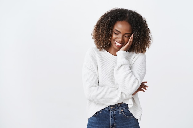 Charmante femme afro-américaine insouciante et insouciante, coiffure frisée, riant, rougissant, regard mignon vers le bas, gloussant, flirty, touche, joue, s'amusant, bonne humeur, plaisantant, conversation humoristique, mur blanc