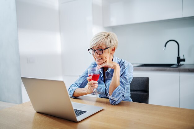 Charmante femme âgée assise dans la cuisine, boire du vin et utiliser un ordinateur portable pour surfer sur Internet