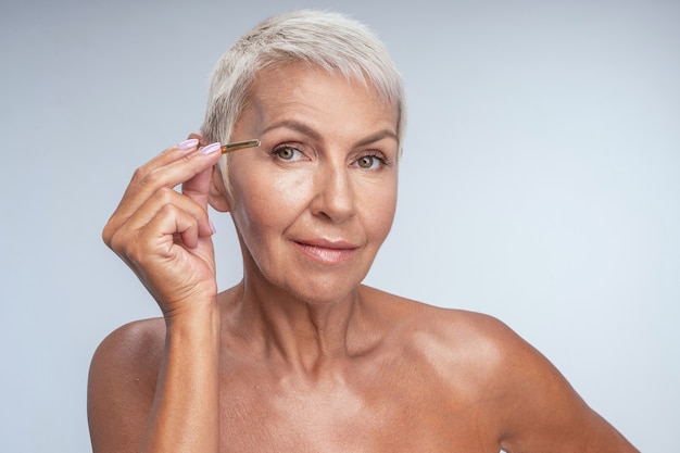 Charmante femme âgée tenant une pince à épiler tout en faisant une nouvelle forme pour les sourcils