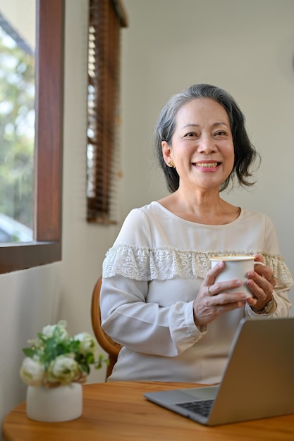 Charmante femme asiatique à la retraite des années 60 tenant une tasse de thé en souriant et en regardant la caméra