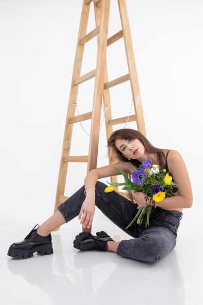 Charmante femme aux cheveux bruns en jeans gris foncé avec bouquet de fleurs violettes et jaunes est assise contre un mur blanc près d'un escalier en bois Concept de beauté féminine et de tendresse