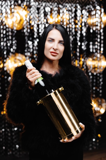 Charmante femme aux cheveux longs brune en manteau de fourrure de soirée à la mode tenant un seau avec une bouteille de champagne sur un mur brillant en studio