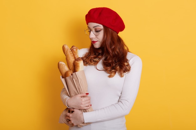 Charmante femme aux cheveux rouges, porte un béret et un pull blanc, regarde un long pain dans sa tête