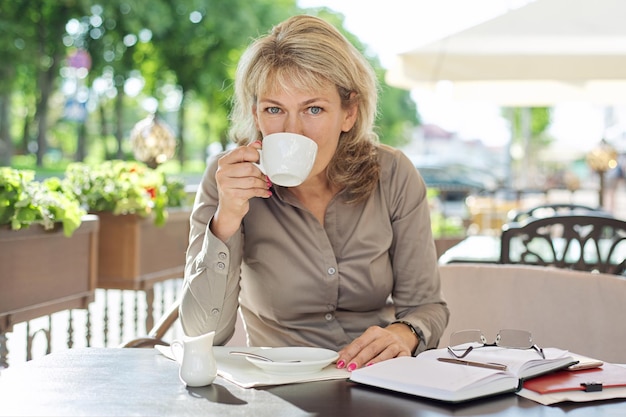 Charmante femme blonde d'âge moyen avec une tasse de café dans un café en plein air, femme d'affaires mature à la pause-café