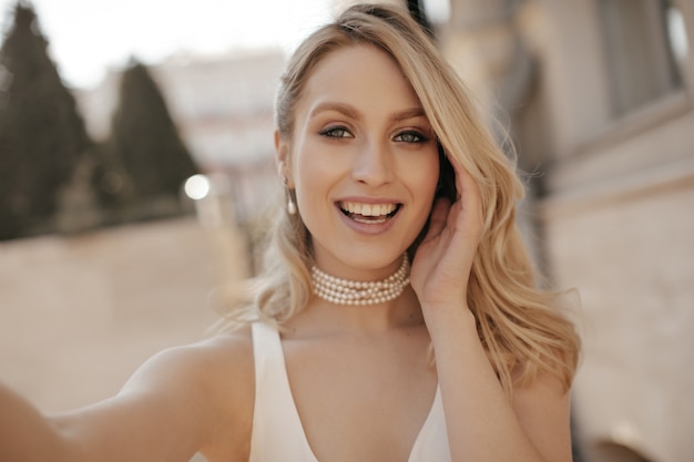 Charmante femme blonde en collier de perles et haut blanc prend un selfie et sourit sincèrement à la rue