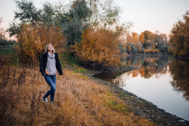 Charmante femme blonde debout au bord de la rivière regardant loin souriant avec des dents avec une veste noire sur les épaules mains sur un pull avec des arbres couverts de coucher de soleil en arrière-plan Promenade d'automne en forêt