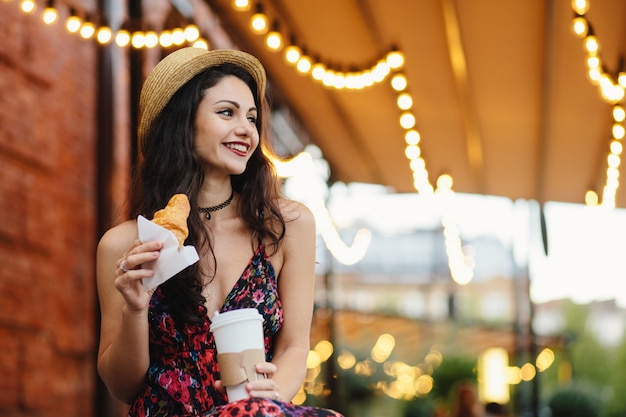 Charmante femme brune avec une apparence agréable portant chapeau et robe d'été tenant un croissant