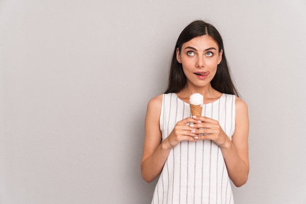 charmante femme brune portant une robe souriante tout en tenant un cornet de crème glacée isolé sur un mur gris