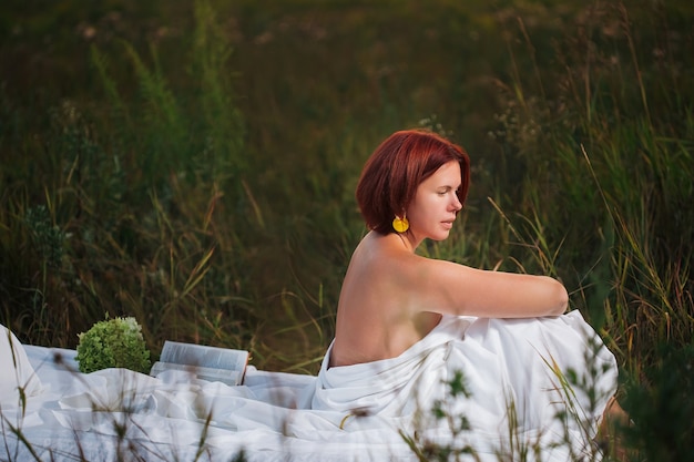 Charmante femme caucasienne assise dans le lit blanc dans un champ au coucher du soleil en été