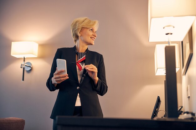 Charmante femme en costume élégant regardant dans le miroir et souriant tout en tenant un téléphone portable et des lunettes