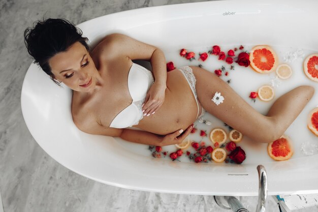Photo charmante femme enceinte souriant tout en se détendant dans une baignoire relaxante avec des fleurs et des huiles essentielles