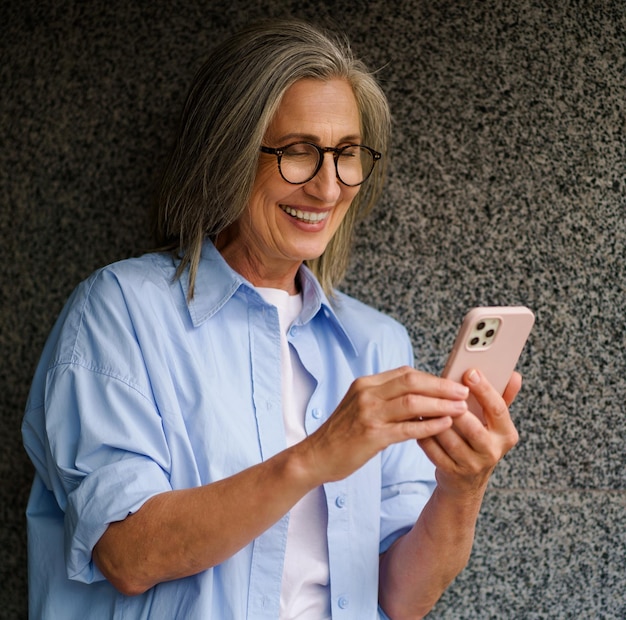 Charmante femme mature aux cheveux gris parlant lors d'un appel vidéo ou lisant un message texte debout à l'extérieur appuyée sur un mur de marbre heureux souriant Debout à l'extérieur positif magnifique femme en chemise bleue décontractée