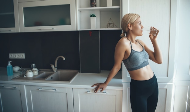 Charmante femme mince en tenue de sport boit de l'eau après avoir fait des exercices de fitness numérique à la maison