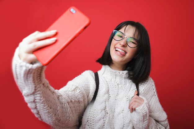 Charmante femme moderne en pull et lunettes prenant selfie et montrant la langue sur fond rouge