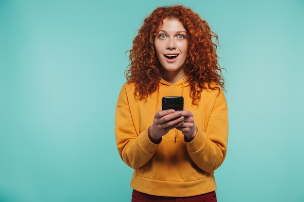 charmante femme rousse des années 20 souriant et utilisant un téléphone portable isolé sur un mur bleu