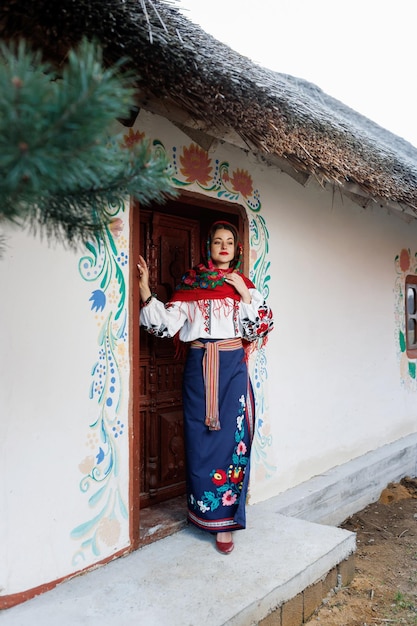 Charmante femme souriante en collier mouchoir ukrainien traditionnel et robe brodée debout à l'arrière-plan d'une cabane décorée Culture ethnique folklorique de style ukrainien