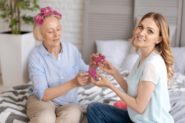 Charmante femme souriante regardant l'avant pendant que sa mère senior s'apprête à enlever les rouleaux de cheveux roses de ses mains