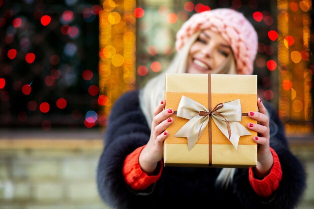 Une charmante fille blonde portant un manteau de fourrure et une casquette vous tire une boîte-cadeau. Espace pour le texte