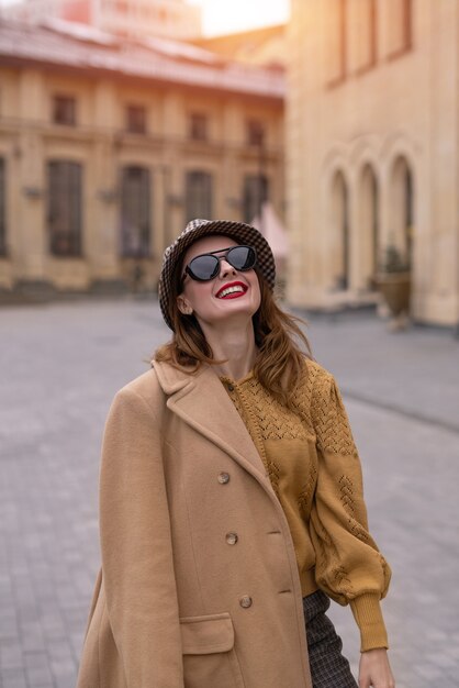 Charmante fille dans un manteau beige d'automne et des lunettes de soleil marchant dans la rue posant joyeusement pour le