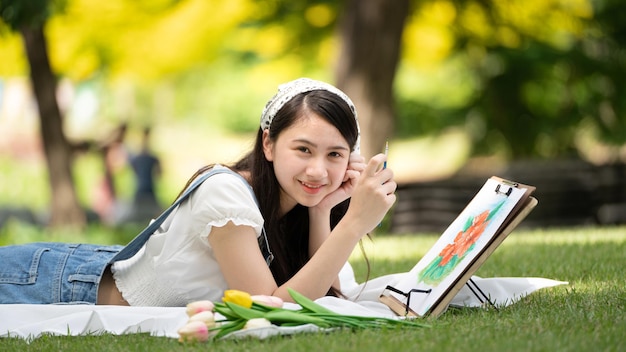 Charmante fille femme assise sur un plaid au parc en journée d'été ensoleillée et à l'aide d'aquarelle