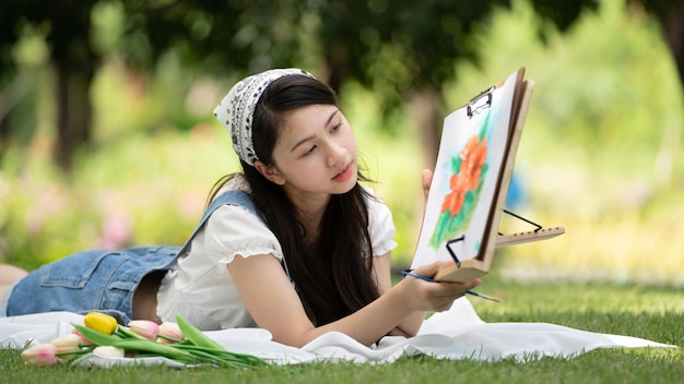 Charmante fille femme assise sur un plaid au parc en journée d'été ensoleillée et à l'aide d'aquarelle