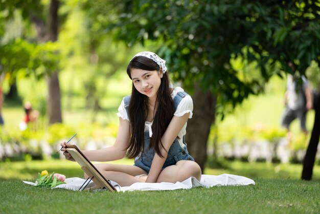 Charmante fille femme assise sur un plaid au parc en journée d'été ensoleillée et à l'aide d'aquarelle