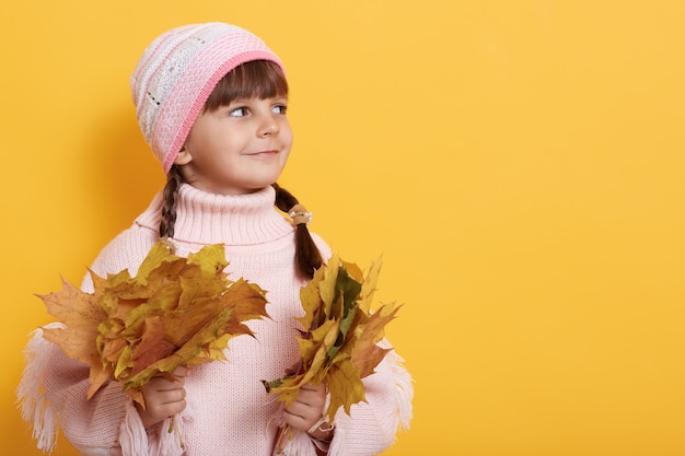 Charmante fille avec des feuilles d'automne à deux mains, enfant regardant de côté avec sourire, posant isolé sur mur jaune