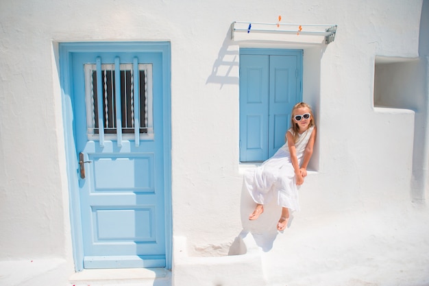Charmante fille en robe blanche à l'extérieur dans les vieilles rues de Mykonos. Enfant dans la rue d'un village traditionnel grec typique avec des murs blancs et des portes colorées sur l'île de Mykonos, en Grèce