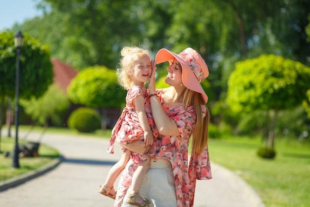 Une charmante fille vêtue d'une robe d'été légère se promène dans un parc verdoyant avec sa petite fille, la tenant dans ses bras.