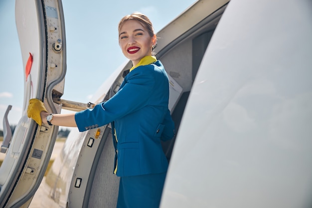 Charmante hôtesse de l'air en uniforme bleu regardant la caméra et souriant tout en plaçant la main sur la porte de l'avion