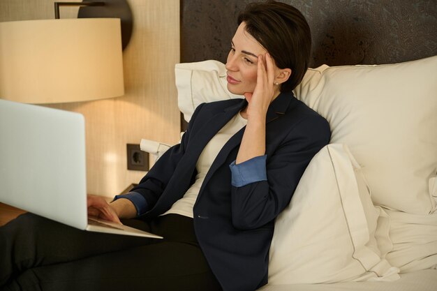 Charmante jeune entrepreneure prospère et pensive, femme d'affaires tenant la main sur le temple tout en tapant sur un ordinateur portable un rapport d'étape, allongée sur un lit dans une chambre d'hôtel lors d'un voyage d'affaires