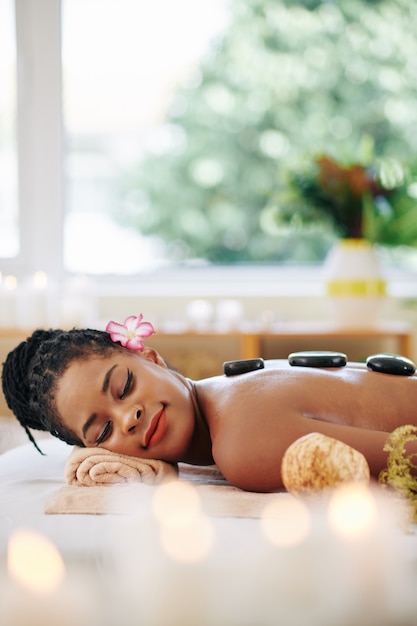Photo charmante jeune femme afro-américaine allongée sur le lit dans un salon de spa avec des pierres de basalte chaudes sur le dos