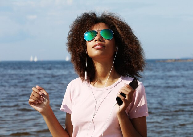 Charmante jeune femme afro-américaine étonnante en lunettes de soleil écoutant de la musique dans des écouteurs sur son téléphone portable