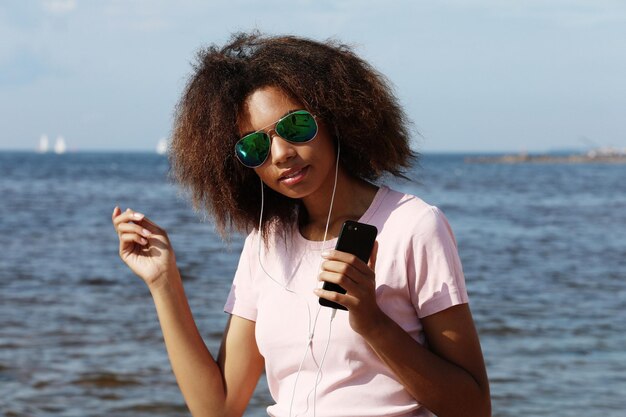 Charmante jeune femme afro-américaine étonnante en lunettes de soleil écoutant de la musique dans des écouteurs sur son téléphone portable