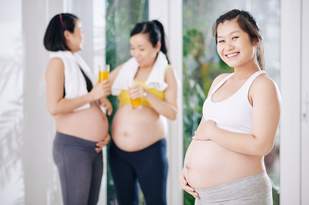 Charmante jeune femme asiatique en haut et leggings touchant le ventre lorsque d'autres femmes enceintes parlent en arrière-plan