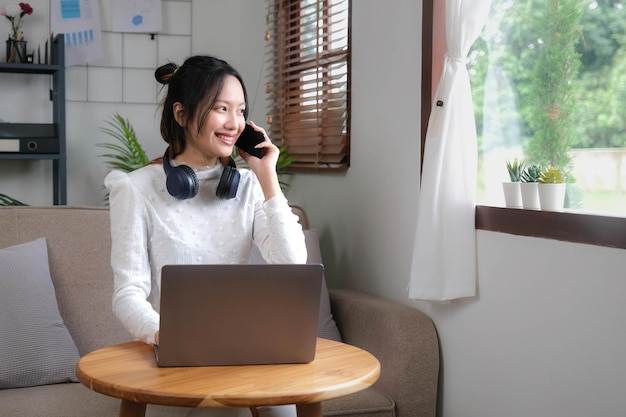 Une charmante jeune femme asiatique se détend dans le salon en appréciant de parler avec son collègue sur son téléphone tout en travaillant à domicile