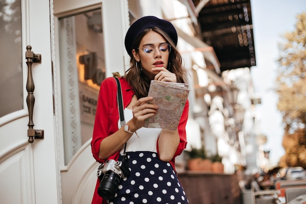 Charmante jeune femme aux cheveux noirs ondulés dans des lunettes et un béret portant une jupe à pois haut blanc chemise rouge caméra sur l'épaule regardant dans la carte sur fond de ville d'automne