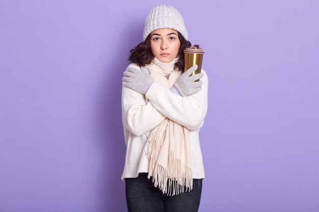 Charmante jeune femme aux cheveux noirs tremblant de froid, essayant de se chauffer, tenant une tasse thermique avec une boisson chaude